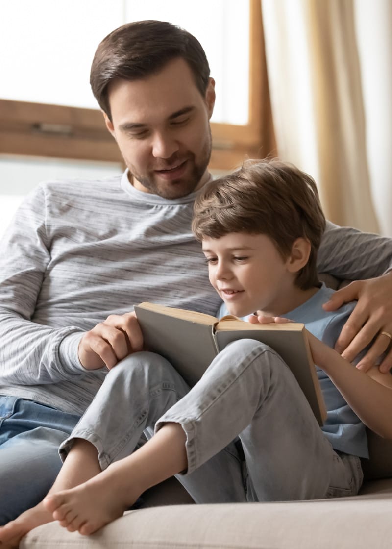 ADHD-Assessment An adult and a child sit together on a couch, reading an open book. The adult has one arm around the child, encouraging learning as they focus on the pages—an ideal setting for informal learning assessments.