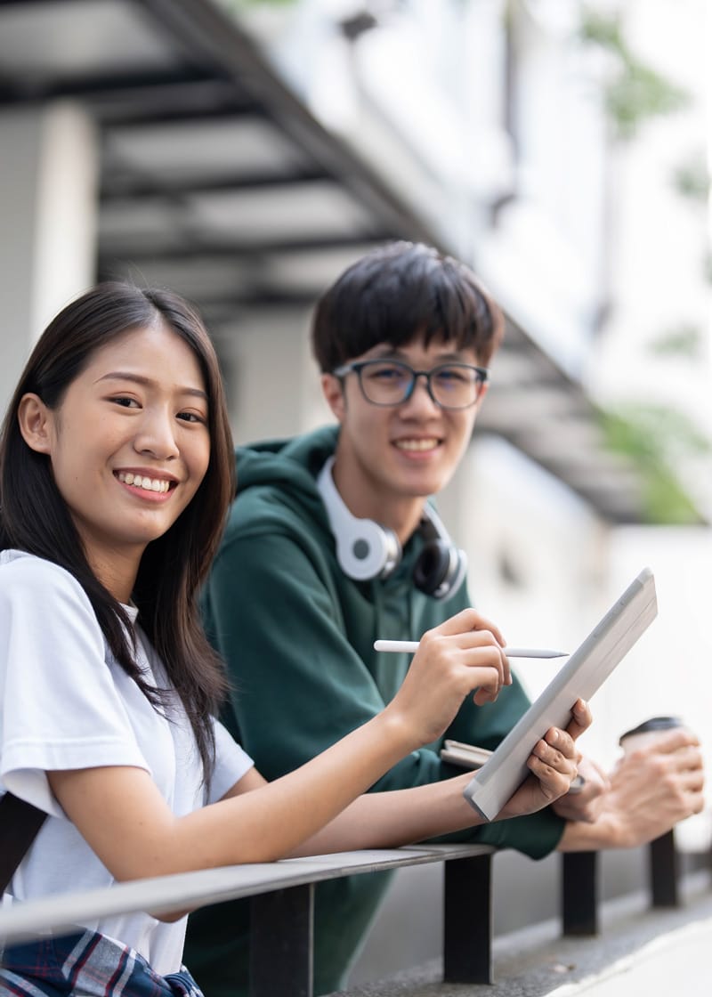 Assessments_1 Two young people standing outdoors, one holding a tablet and stylus, both smiling at the camera. The scene suggests a positive experience, possibly related to learning or cognitive assessments. A building with blurred details forms the background.