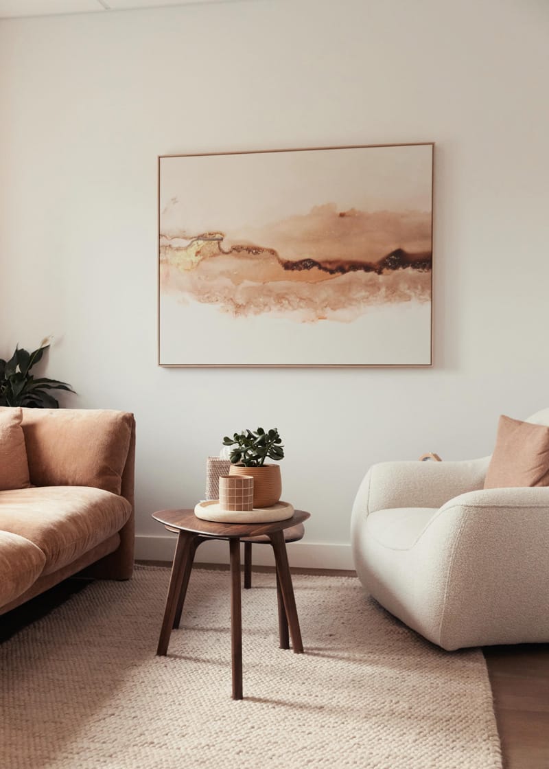 A modern living room with a tan sofa, cream armchair, wooden side table with decor, beige rug, and abstract art on the wall.