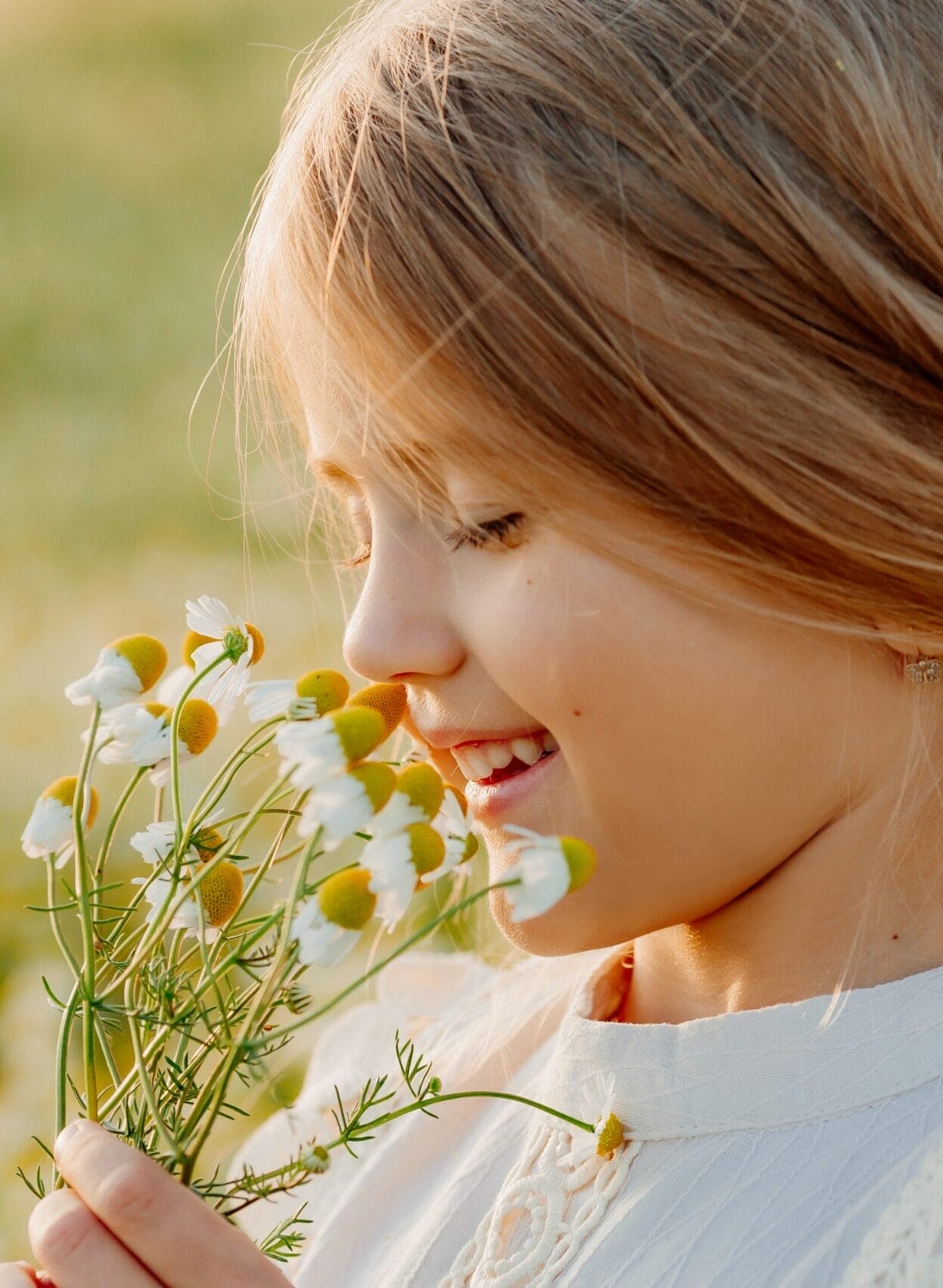 A young girl with light brown hair in a braid smiles while smelling a bunch of small white and yellow flowers outdoors.