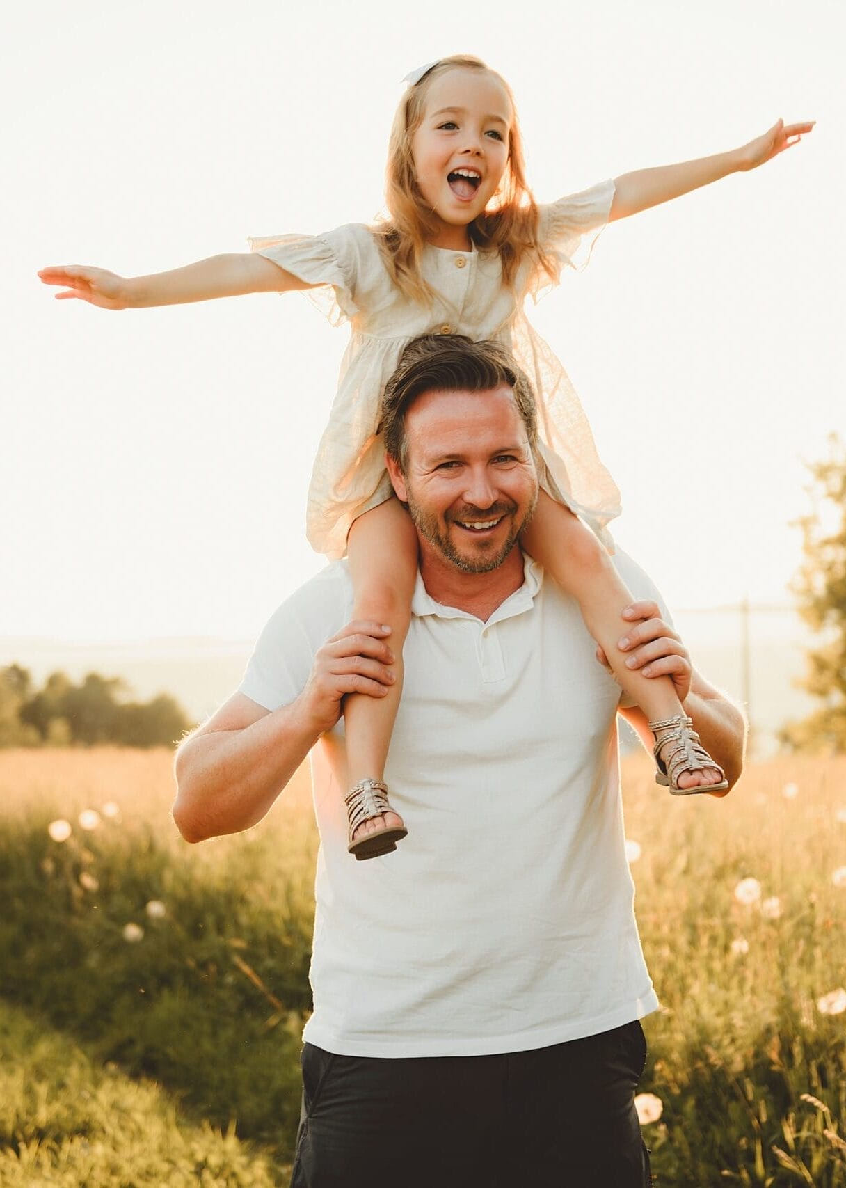 A man stands in a sunny field smiling with a young girl sitting on his shoulders, her arms outstretched.